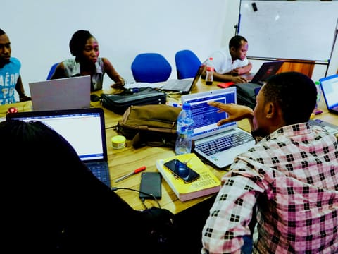 A group of learners work on laptops around a shared table, one pointing at a screen to explain a step