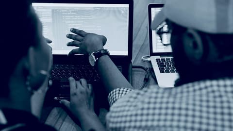 Hands typing on a laptop showing code, with a colleague working alongside